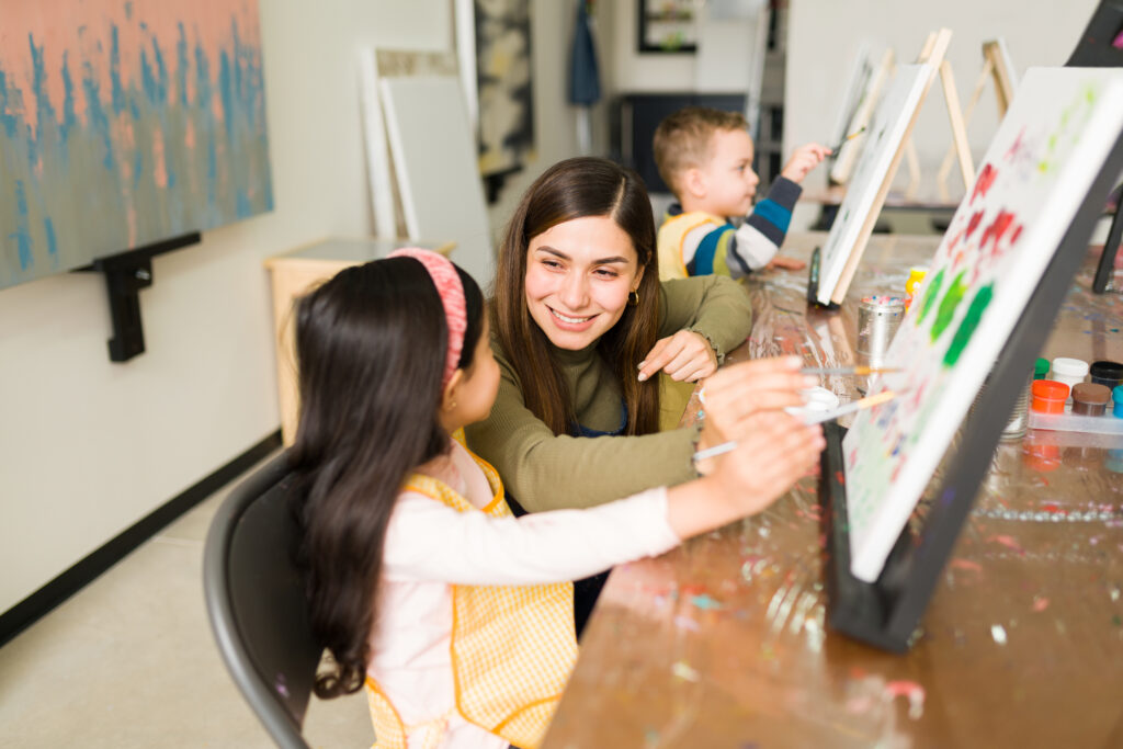 Hispanic female instructor smiling and teaching a little girl how to paint on a canvas during a painting class for children | Family Night | Merely Creative | Martinsburg WV