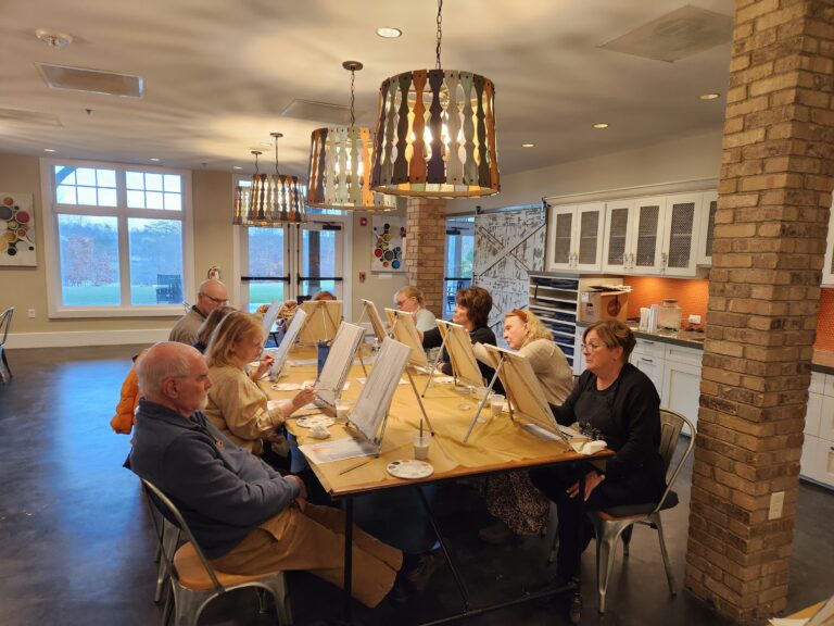 Group sitting at a table with canvases, easels, and painting supplies to paint a picture | Wine and Paint Night | Merely Creative Martinsburg WV
